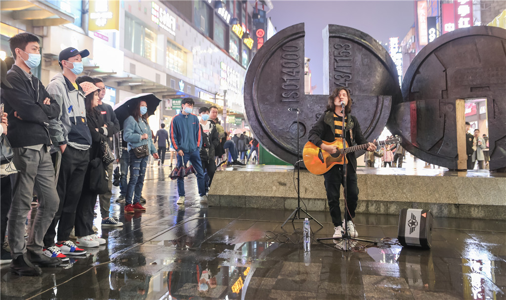 2021年4月13日晚，湖南省长沙市，雨中的黄兴路步行街上，市民和游客在观看街头歌手的演唱。